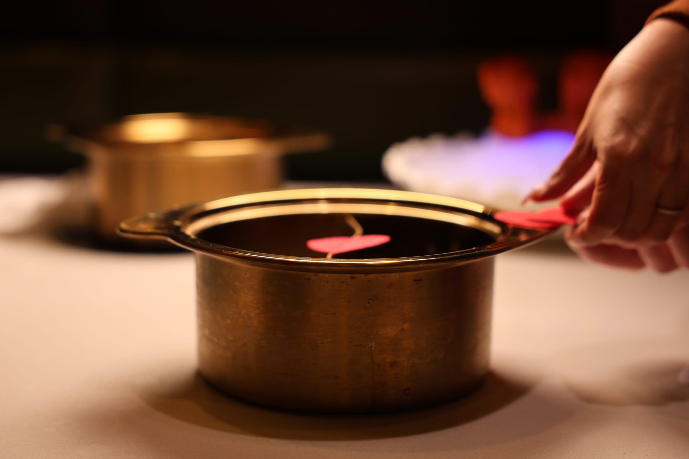 A brass hot pot being set down on a dark table, steam catching the light.
