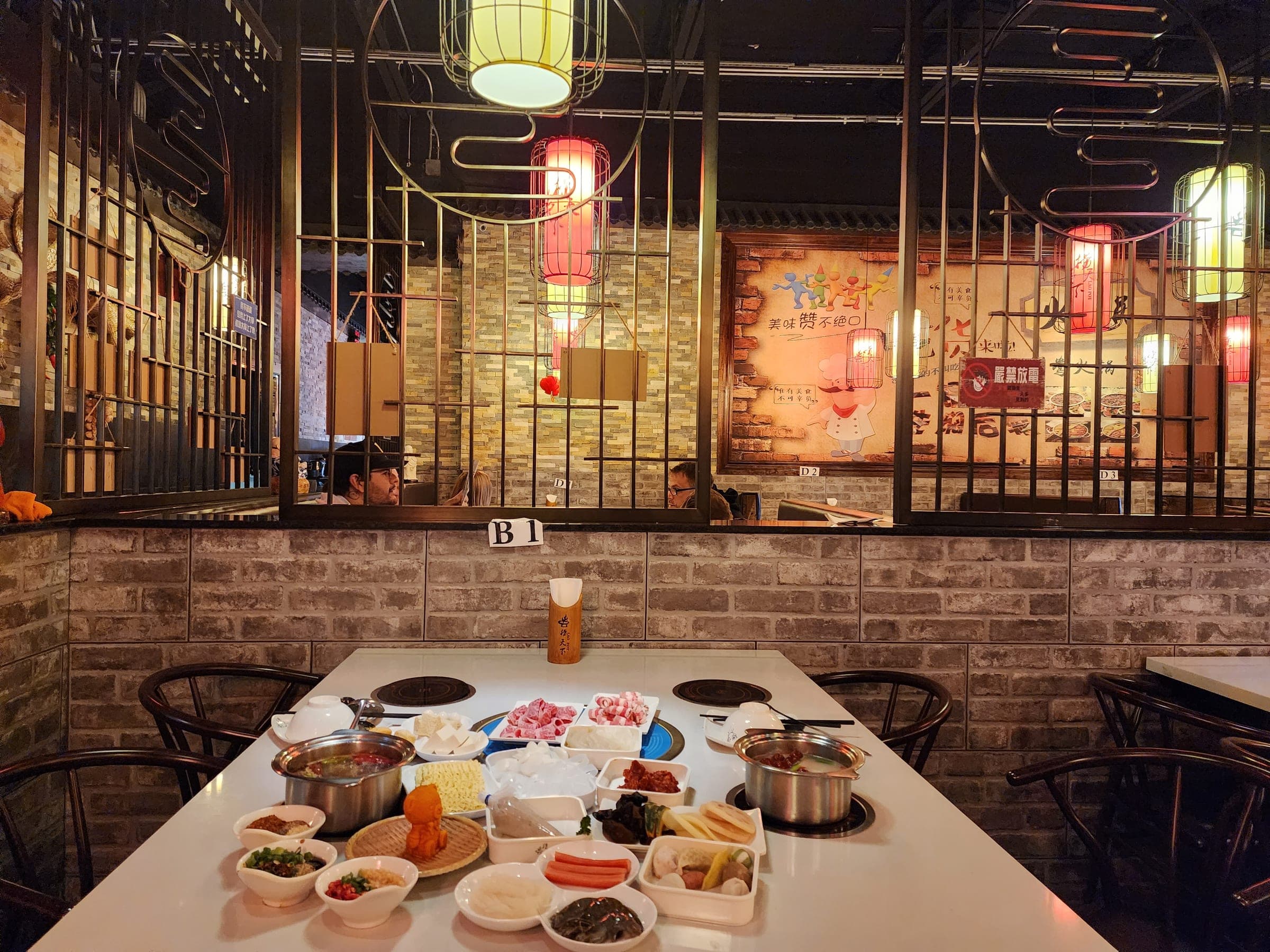 A long white table set with two hot pots and a dozen small dishes, under red and cream paper lanterns beside a brick half-wall.